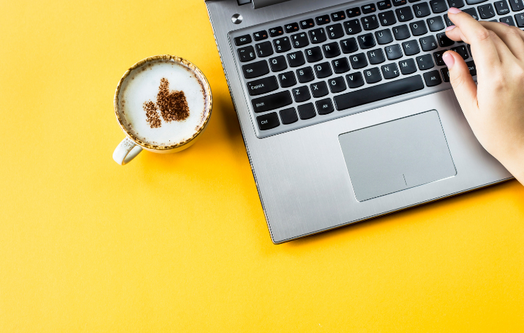 A hand typing on a laptop beside a coffee cup with a thumbs up design on top