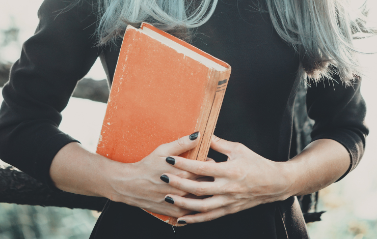 A witchy person in a black dress holding an orange book