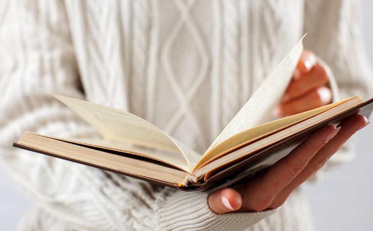A torso covered in a white knit sweater holding open a book