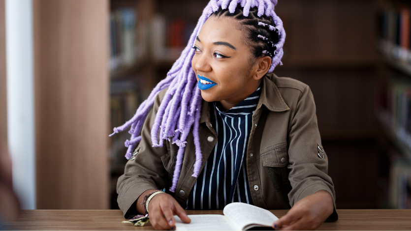 A young black woman at a library with an open book looking out the window and smiling