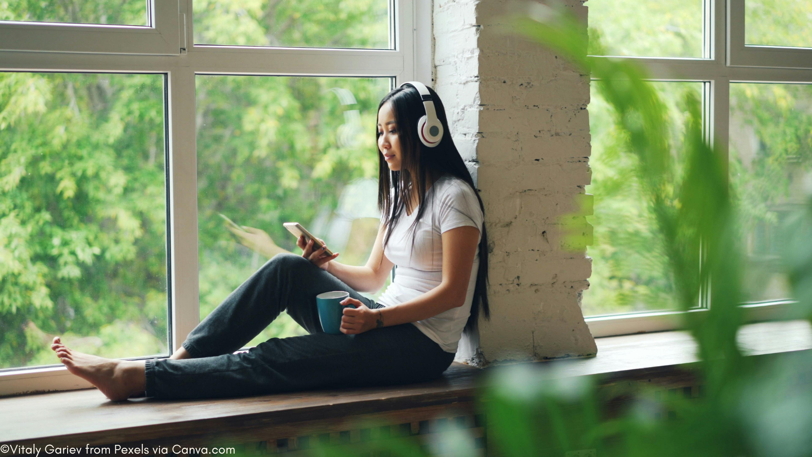 A woman sitting on a window ledge, wearing headphones, and holding a phone