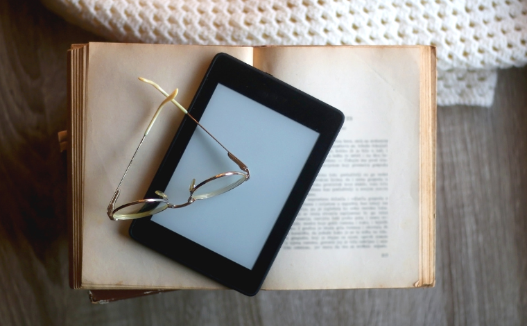 An e-reader sitting on an open book beneath a pair of gold glasses