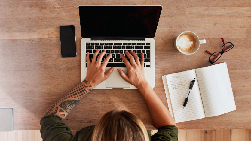 Above shot of someone with their hands on a laptop keyboard. A phone is to their left, to their right is an open note book, coffee, and glasses.