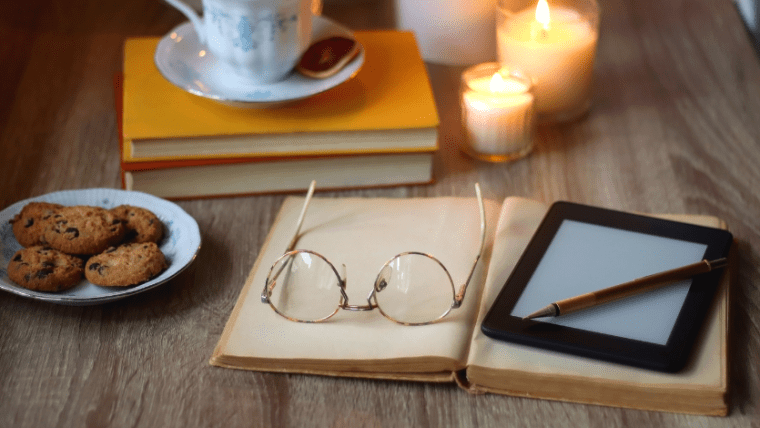 Glasses and an e-reader on top of an open book with books and candles in the background