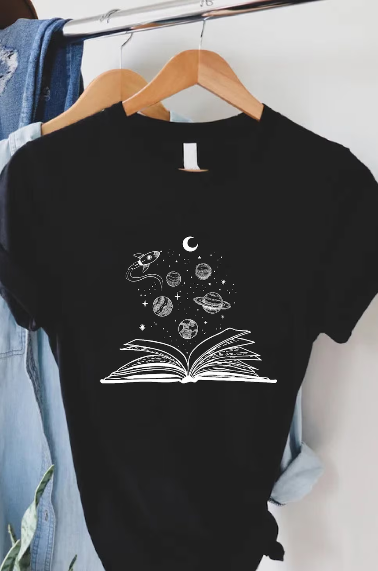 A black t-shirt with a white design of an open book and planets and stars floating out from the center of the book