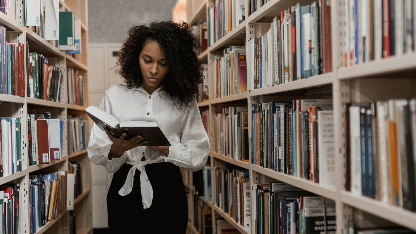 A Black woman looking at a book between bookshelves